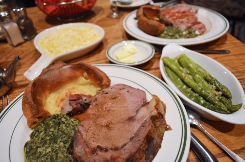 A table setting with plates of prime rib, Yorkshire pudding, spinach, asparagus, and a side of butter.