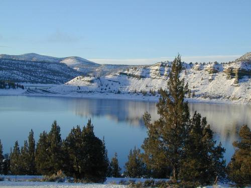 A serene winter landscape featuring a calm lake, snow-covered hills, and evergreen trees under a clear blue sky.