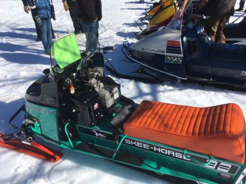 A vintage snowmobile with a green and orange seat parked on snow, surrounded by other snowmobiles and people.