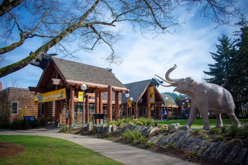 A wooden building with a large elephant statue in front, surrounded by trees and a pathway.