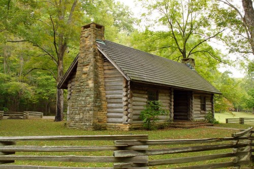A rustic log cabin with a stone chimney, surrounded by trees and a wooden fence in a serene outdoor setting.