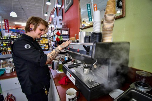 Barista with tattoos prepares coffee at an espresso machine, steam rising in a cozy café setting.
