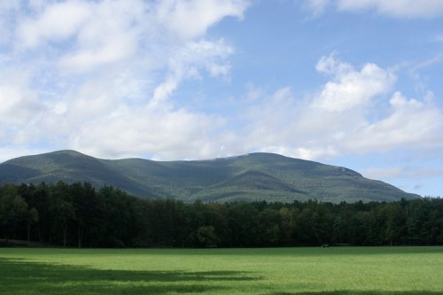 A lush green field in the foreground with a backdrop of rolling mountains under a partly cloudy sky.