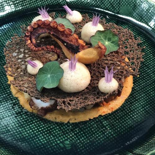 A gourmet dish featuring grilled octopus on a textured plate, garnished with cream spheres and edible flowers.