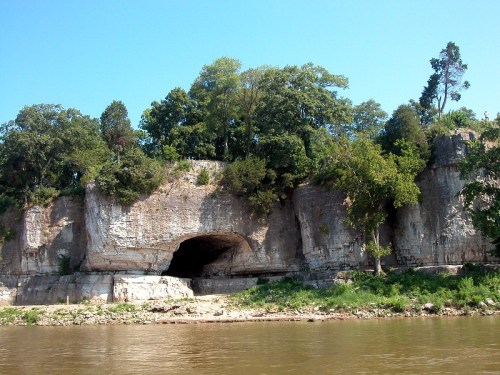 A rocky cliff with a large cave entrance, surrounded by lush green trees and reflecting in a calm river below.