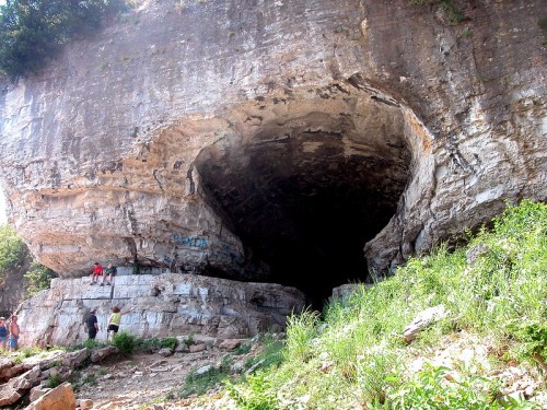 A large cave entrance with rocky walls, surrounded by greenery and people exploring the area.