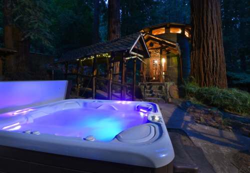 A hot tub with blue lights in a forest setting, near a cozy cabin surrounded by tall trees at dusk.