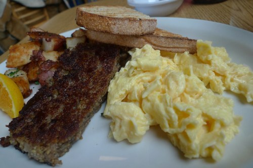 A plate featuring scrambled eggs, meatloaf, toast, and crispy potatoes, garnished with a slice of orange.