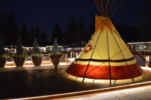 A brightly colored teepee illuminated at night, surrounded by trees and a wooden deck with potted plants.