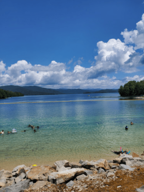 A serene lake with clear blue water, surrounded by trees and mountains, with people swimming and enjoying the sun.