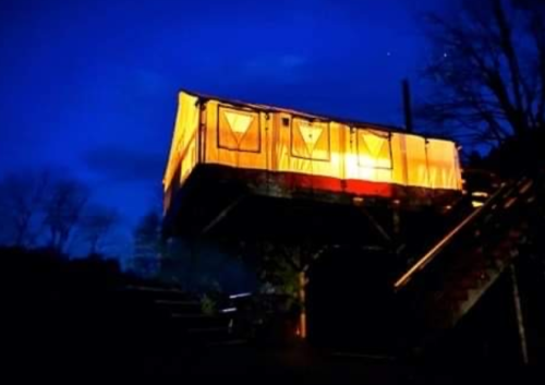 A brightly lit cabin on stilts at dusk, surrounded by trees and a dark blue sky.