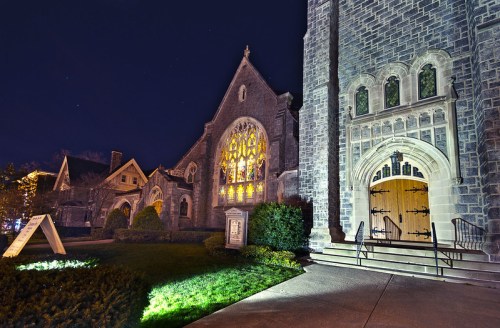 A stone church illuminated at night, featuring a large stained glass window and a welcoming entrance.