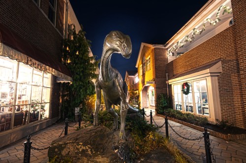 A dinosaur statue stands in a well-lit outdoor shopping area at night, surrounded by festive decorations and brick buildings.