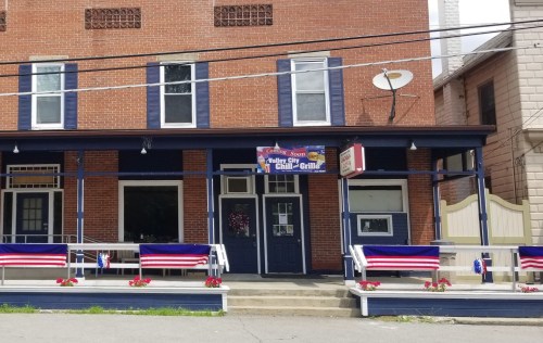 A brick building with blue trim, featuring a sign for a chili grill and patriotic decorations on the porch.