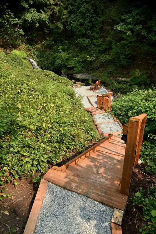A wooden staircase leads down through lush greenery to a small waterfall and rocky area.
