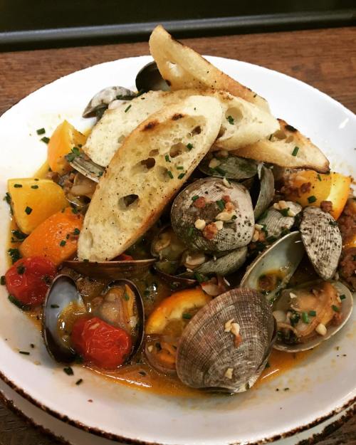 A bowl of seafood stew with clams, mussels, colorful vegetables, and toasted bread on top, garnished with herbs.