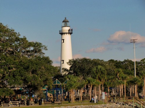 A tall white lighthouse stands near a park with palm trees and playground equipment under a clear blue sky.