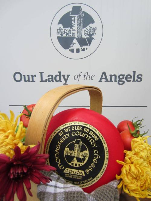 A round wheel of Gouda cheese in a basket, surrounded by flowers and tomatoes, with a sign reading "Our Lady of the Angels."