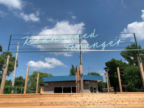 Neon sign reading "Redheaded Stranger" against a blue sky with clouds, framed by trees and a building in the background.