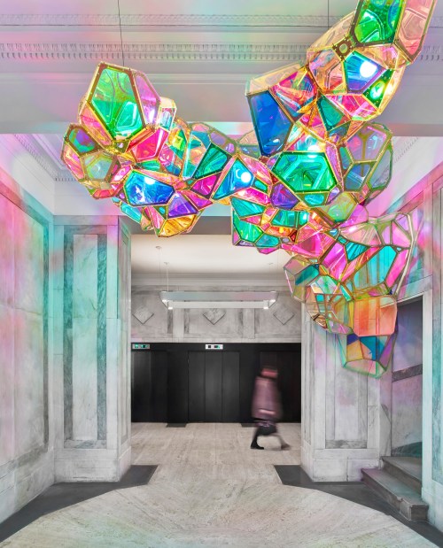 A colorful, abstract light installation hangs from the ceiling in a marble hallway, with a blurred figure walking by.
