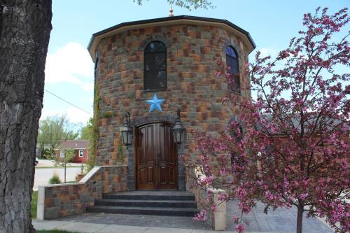 A stone building with a round tower, wooden doors, and a blue star, surrounded by blooming trees and a clear sky.