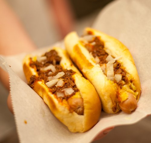 Two chili hot dogs topped with onions, held in a paper wrapper.