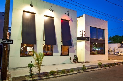 Modern restaurant exterior at dusk, featuring large windows, black awnings, and a welcoming entrance.