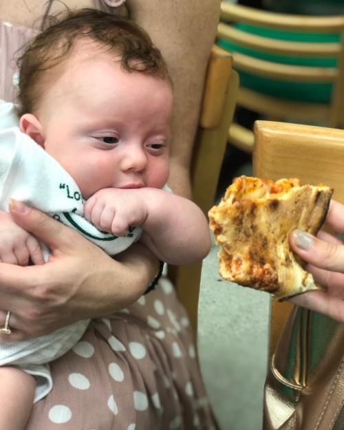 A baby curiously reaches for a slice of pizza held by an adult, sitting in a casual dining setting.