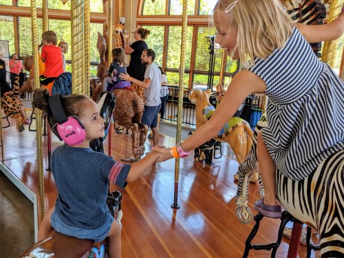 Two children on a carousel, one wearing headphones, reaching out to each other while surrounded by animal figures.
