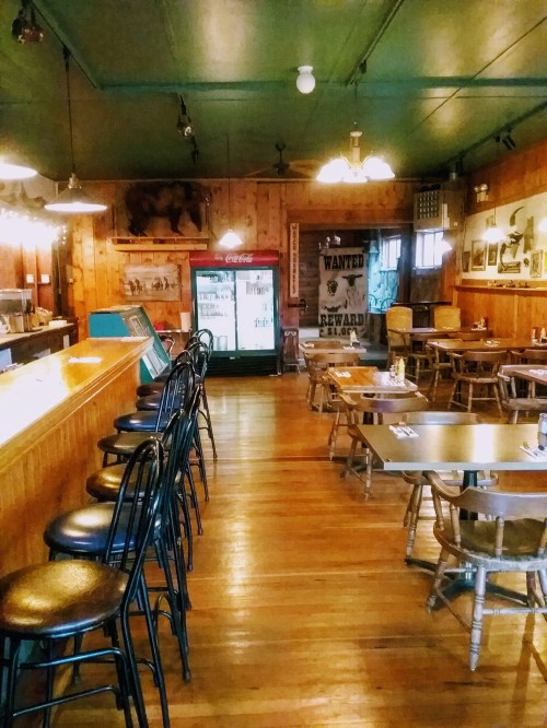 A rustic interior of a restaurant with wooden walls, tables, and a bar, featuring a vintage Coca-Cola fridge.