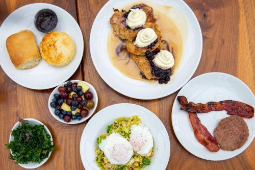 A spread of breakfast dishes including pancakes with blueberries, bacon, eggs, a biscuit, and a fruit bowl.