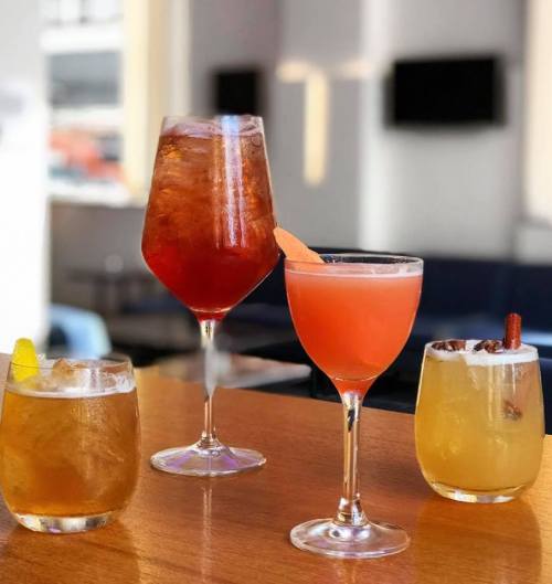 Four colorful cocktails on a wooden table, featuring a mix of garnishes and glass styles in a bright setting.