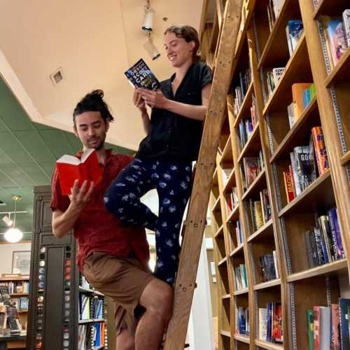 Two people in a bookstore: one on a ladder reading, the other holding a book and looking up, surrounded by shelves.