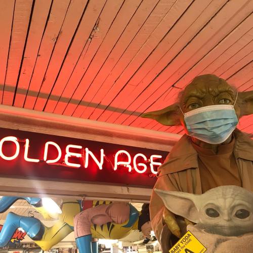 A small, green character wearing a mask stands under a sign that reads "Golden Age," surrounded by colorful decorations.