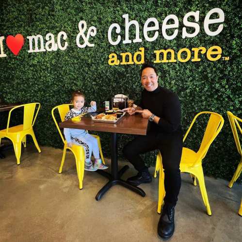 A woman and a child sit at a table in a restaurant with a green wall that says "I ❤️ mac & cheese and more."