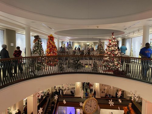 A festive display of decorated Christmas trees in a multi-level atrium, with visitors admiring the decorations.