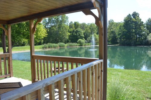 A wooden gazebo overlooks a serene pond with a fountain, surrounded by lush greenery and trees under a clear blue sky.