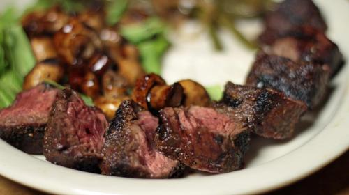 A plate of grilled steak cubes, sautéed mushrooms, and greens, served with a side of rice.