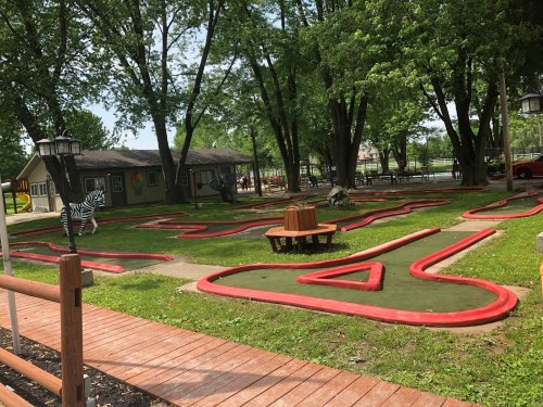 A mini-golf course with red borders, surrounded by trees and a small building in the background.