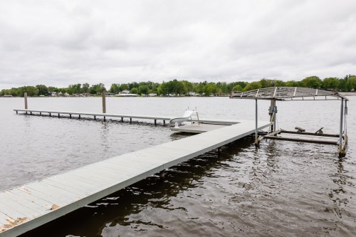 A wooden dock extends into a calm lake, with a covered boat lift and trees in the background under a cloudy sky.
