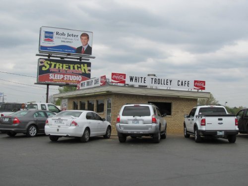 A small café with a sign reading "White Trolley Café," surrounded by parked cars and a billboard in the background.