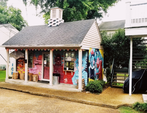 A colorful, mural-covered ice cream stand with a porch and decorative lights, surrounded by greenery.
