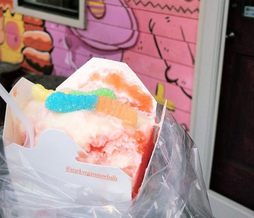 A colorful snowball dessert topped with gummy worms, held in a takeout container against a vibrant mural background.