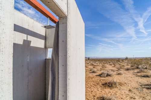 A minimalist outdoor shower structure made of concrete, set against a desert landscape under a blue sky.