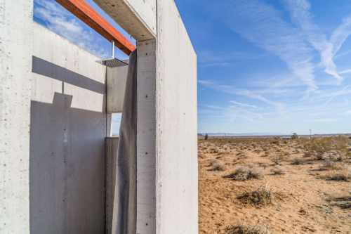 A minimalist outdoor shower structure made of concrete, set against a desert landscape under a blue sky.