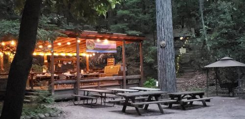 A rustic outdoor gathering space with picnic tables, string lights, and a wooden pavilion surrounded by trees.