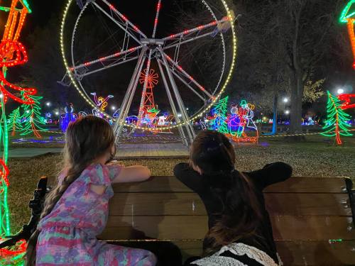 Two children sit on a bench, watching a brightly lit Ferris wheel and festive decorations at night.