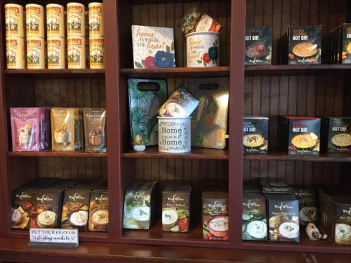 A wooden shelf displays various food products, including soups, dips, and snacks, with decorative containers and signs.