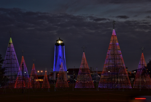 Colorful illuminated Christmas trees in a festive display, with a lighthouse glowing in blue in the background at dusk.