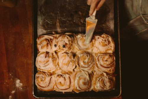 A hand icing freshly baked cinnamon rolls on a baking tray, with a wooden surface in the background.